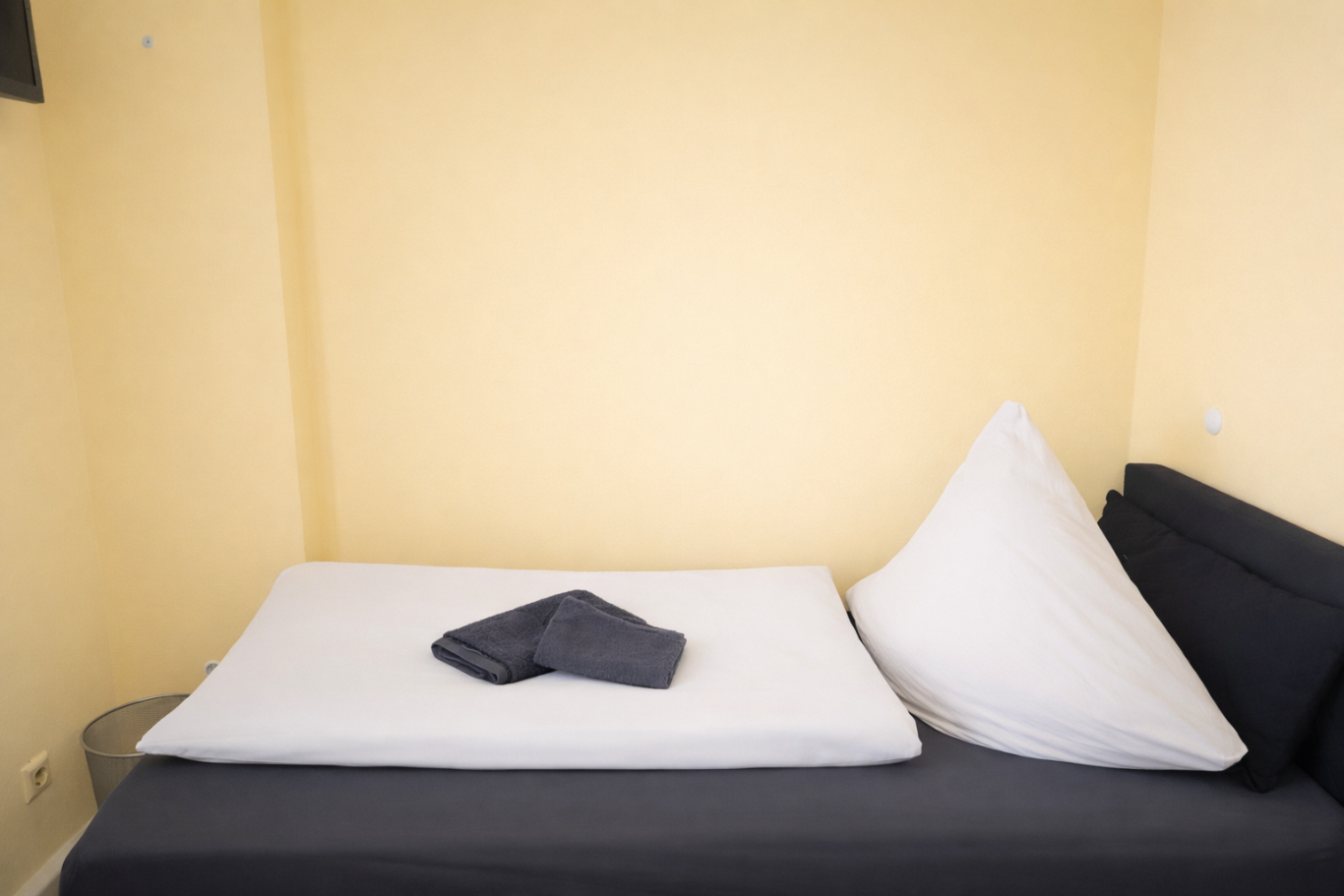 Ein einfaches Bett mit einer weißen Matratze und einem grauen Handtuch auf gelbem Hintergrund.