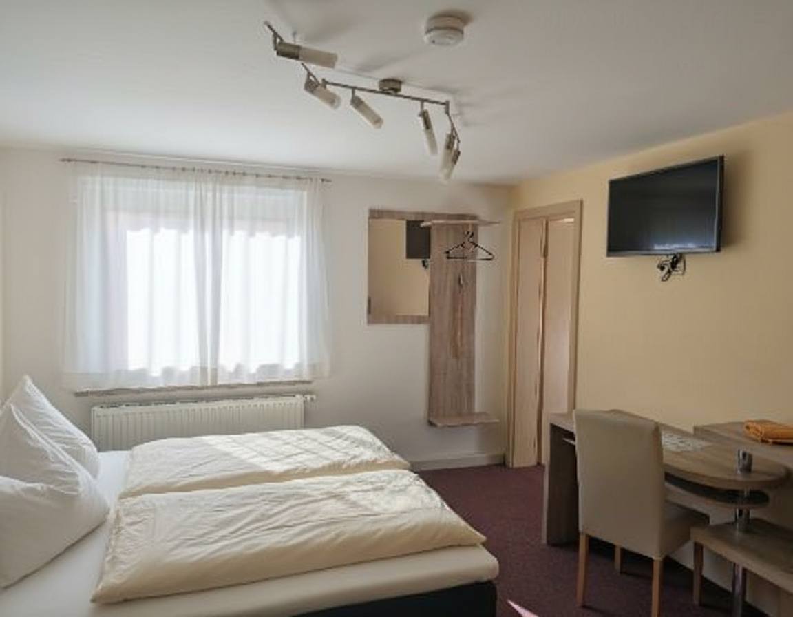 Helles Zimmer mit zwei Betten, Schreibtisch, Fernseher und einem Fenster mit Vorhängen.