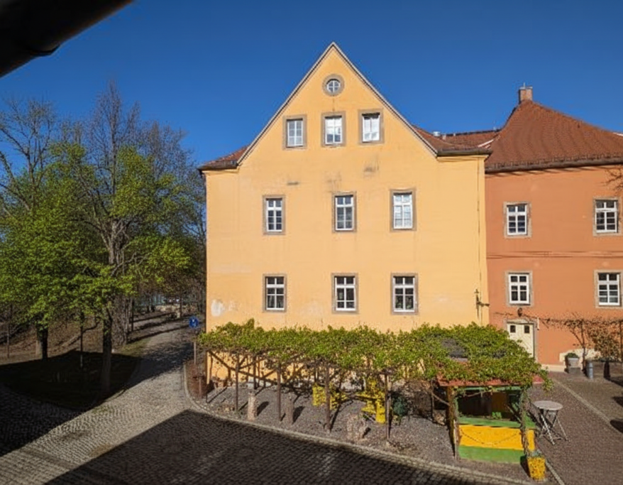 Gelbes Gebäude mit Fenster und angrenzender grüner Fläche bei klarem Himmel.